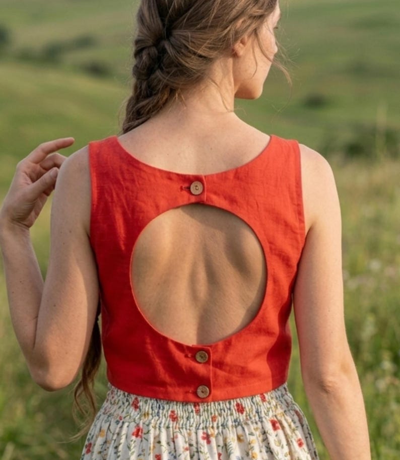 May include: A sleeveless, cropped red top featuring a large circular cutout on the back. The garment includes three small, round, light brown buttons positioned along the center back, with one at the top of the cutout and two at the bottom hem.