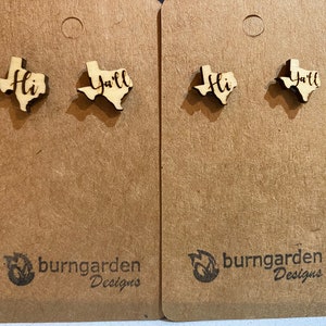 May include: Two pairs of wooden earrings shaped like the state of Texas. One pair says "Hi" and the other pair says "Y'all". The earrings are displayed on a brown card with the text "burngarden Designs".