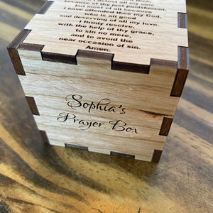 May include: A wooden prayer box with a light brown finish and dark brown trim. The box has an inscription that reads "Sophia's Prayer Box" and a prayer titled "Act of Contrition".