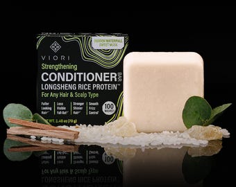 May include: A rectangular off-white conditioner bar next to its black packaging box labeled Viori Strengthening Conditioner Bar Longsheng Rice Protein. The box displays 2.48 oz (70 g) and 100 washes. White rice grains, two green leaves, and two wooden sticks are placed in front.