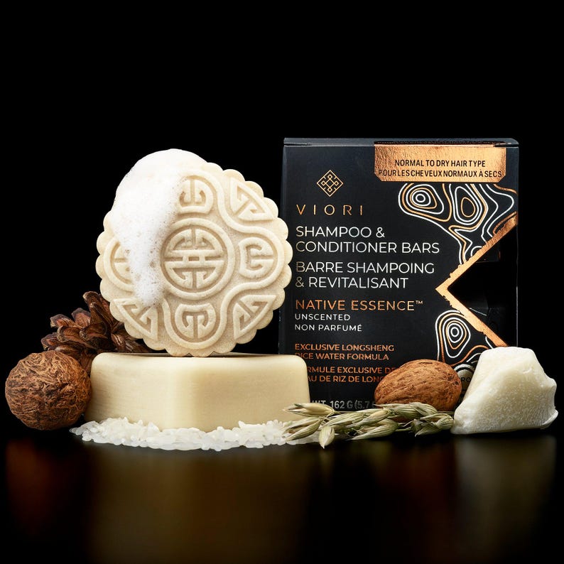 May include: A Viori shampoo and conditioner bar set featuring a round, embossed bar with white foam, a rectangular base bar, and a black box labeled Native Essence. The set includes raw rice grains, a pinecone, a nut, and oat stalks as accents.
