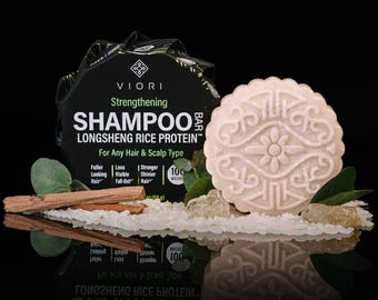 May include: A round, beige shampoo bar with an embossed floral pattern sits next to its black packaging. The box reads Viori Strengthening Shampoo Bar Longsheng Rice Protein for Any Hair & Scalp Type, 3.1 oz (88 g). White rice grains and wooden sticks are visible.