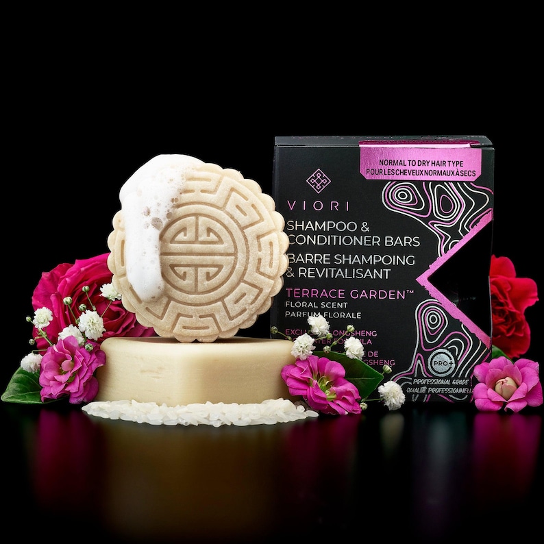 May include: A round, beige shampoo bar with a geometric embossed pattern and white foam, resting on a rectangular base. Beside it is a black box labeled Viori Shampoo & Conditioner Bars Terrace Garden, surrounded by pink roses and white baby's breath.
