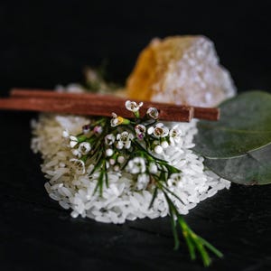 May include: A cluster of white waxflower blossoms with green stems rests atop a pile of white rice grains. Two brown wooden sticks lie horizontally across the rice, accompanied by a translucent amber-colored crystal and a single green leaf.