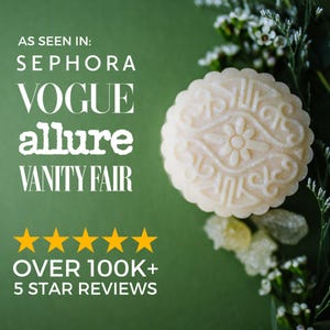 May include: A round, off-white soap bar with an embossed floral pattern. Text reads: AS SEEN IN: SEPHORA VOGUE allure VANITY FAIR. Five yellow stars are displayed above the text: OVER 100K+ 5 STAR REVIEWS. The soap is set against a dark green background.