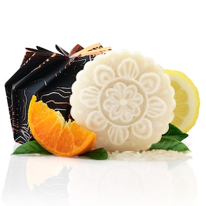 May include: A white, round soap bar with a floral design, a slice of orange, a lemon, and a black box with orange and white stripes. The soap bar is surrounded by white rice.