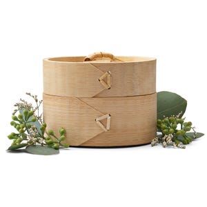 May include: A two-tiered round bamboo steamer with a natural wood finish. The steamer has a woven bamboo handle and is sitting on a white surface with green leaves.