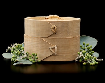 May include: A two-tiered bamboo steamer with a light tan wood grain finish and a woven lid handle. The cylindrical container features triangular bamboo reinforcements on the sides and is surrounded by sprigs of green eucalyptus leaves.