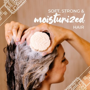 May include: A round, off-white shampoo bar with an embossed pattern being used to lather wet hair. The text SOFT, STRONG & moisturized HAIR is printed in white above the hair. The corners feature a white geometric line art design.