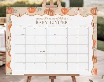 May include: A large white calendar on a wooden easel, with a border of pumpkins and greenery. The text reads "Guess the arrival date for BABY JUNIPER." The calendar grid is blank, with dates for April, May, and June. The easel is made of light brown wood.