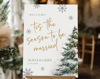 May include: A white sign with gold lettering that reads "Tis the season to be married" with a watercolor illustration of a pine tree. The sign also includes the names Olivia & Caden and the date 12.16.20XX. The sign is on a wooden easel.