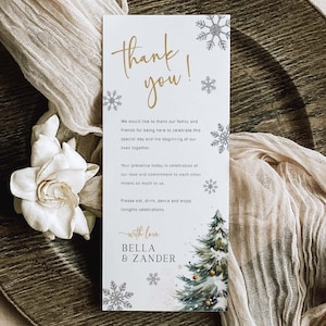 Puede incluir: Una tarjeta de agradecimiento de boda con la inscripción dorada "thank you!" y un mensaje. La tarjeta tiene ilustraciones de copos de nieve y árboles de Navidad. La tarjeta está sobre un plato de madera con una flor blanca y una tela beige.