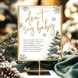 Puede incluir: Un letrero con marco dorado con las palabras "don't say baby" en escritura elegante. El letrero incluye instrucciones para un juego de baby shower, con ilustraciones de copos de nieve y árboles de Navidad. El letrero se muestra en un soporte dorado.