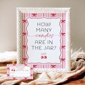 Puede incluir: Un letrero rosa y blanco enmarcado con el texto "HOW MANY candies ARE IN THE JAR?" y una tarjeta más pequeña a juego. El letrero tiene un borde decorativo con lazos rojos y cerezas. Los artículos están en una bandeja de madera.