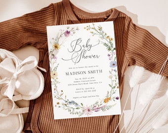 May include: A baby shower invitation with a floral wreath design, featuring the text "Baby Shower" and event details for Madison Smith. The invitation rests on a brown ribbed babygrow, with white baby booties and dried flowers.