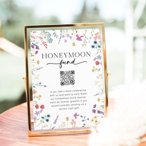 May include: A white sign with a floral border and the text "HONEYMOON fund" with a QR code. The sign is in a gold frame and is sitting on a wooden table.