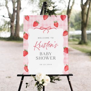 May include: A pink and white gingham baby shower welcome sign with red strawberries and a red ribbon. The sign reads "Welcome to Kristen's Baby Shower 08.20.20XX".