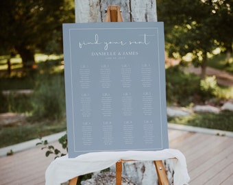 Pode incluir: Um diagrama de lugares de casamento azul com texto branco. O diagrama é intitulado "find your seat" e lista os nomes dos convidados sob diferentes números de mesa. O diagrama está em um cavalete de madeira.