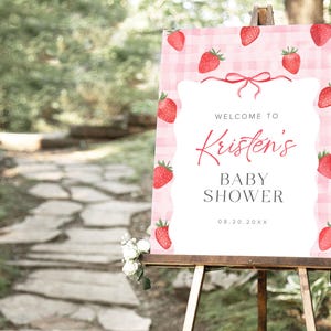 May include: A pink and white gingham baby shower welcome sign with red strawberries and the text "Welcome to Kristen's Baby Shower 08.20.20XX".