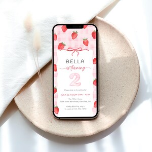 May include: A smartphone displays a birthday invitation with a strawberry theme. The invitation features the text "BELLA is turning 2" on a pink gingham background, with strawberry illustrations. The phone rests on a beige plate.