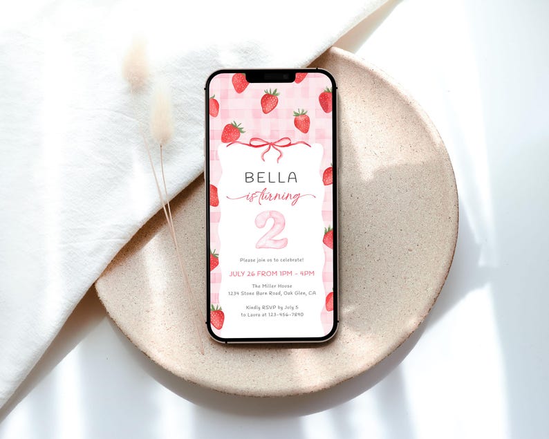 May include: A smartphone displays a birthday invitation with a strawberry theme. The invitation features the text "BELLA is turning 2" on a pink gingham background, with strawberry illustrations. The phone rests on a beige plate.