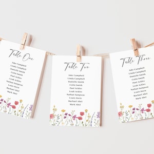 Könnte beinhalten: Drei weiße Karten mit floralen Rändern und schwarzem Text. Die Karten sind mit "Tisch Eins", "Tisch Zwei" und "Tisch Drei" beschriftet und listen die Namen für jeden Tisch auf.