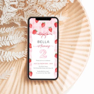 Może przedstawiać: Smartfon wyświetla zaproszenie urodzinowe z motywem truskawek. Zaproszenie brzmi "BELLA is turning 2" z datą i godziną przyjęcia. Telefon spoczywa na ozdobnym talerzu.