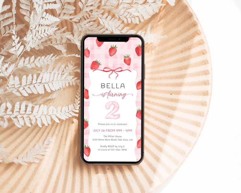 Może przedstawiać: Smartfon wyświetla zaproszenie urodzinowe z motywem truskawek. Zaproszenie brzmi "BELLA is turning 2" z datą i godziną przyjęcia. Telefon spoczywa na ozdobnym talerzu.