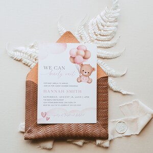 Pode incluir: Um convite de ch&aacute; de beb&ecirc; rosa e marrom com um fundo de aquarela. O convite apresenta um urso de pel&uacute;cia segurando bal&otilde;es e o texto "We can barely wait" para a beb&ecirc; Amelia. O convite inclui a data, a hora e o local do ch&aacute; de beb&ecirc;.