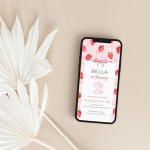 May include: A smartphone displays a birthday invitation with a strawberry theme. The invitation features the name "Bella" and the number "2" in pink, surrounded by strawberry illustrations. White palm leaves are in the background.