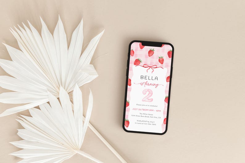 May include: A smartphone displays a birthday invitation with a strawberry theme. The invitation features the name "Bella" and the number "2" in pink, surrounded by strawberry illustrations. White palm leaves are in the background.