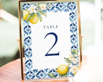 Puede incluir: Número de mesa dos con un borde de azulejos azules y blancos y acentos de limón y flores. La tarjeta está en un marco dorado.