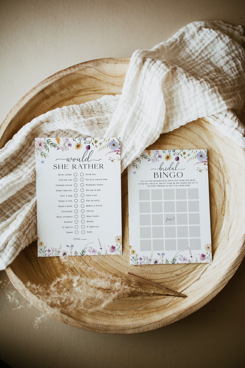May include: Two floral-themed cards for a bridal shower. One card is a "Would She Rather" game with a list of questions and checkboxes. The other card is a bridal bingo card with a floral border.