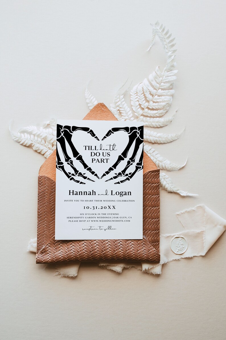 Puede incluir: Una invitaci&oacute;n de boda blanca con manos de esqueleto negras formando una forma de coraz&oacute;n. El texto dice "Till Death Do Us Part" e incluye los nombres Hannah y Logan, la fecha 10.31.20XX y la ubicaci&oacute;n Serendipity Garden Weddings | Oak Glen, CA.