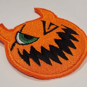 May include: Embroidered orange patch in the shape of a jack-o'-lantern with black jagged teeth and a stitched-on green eye. The patch has two small horns and a stitched border.