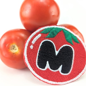 Puede incluir: Tres tomates rojos maduros y un parche con forma de tomate. El parche es rojo con un contorno blanco, hojas verdes y la letra negra "M" en el centro. El fondo es blanco.