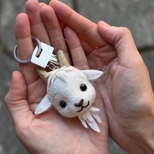 May include: A white felt goat head keychain featuring small tan horns, black bead eyes, and embroidered facial details including a nose, mouth, and a pointed beard. The goat head is attached to a silver-toned metal split ring key holder.