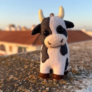 Może przedstawiać: Mała filcowa figurka krowy w biało-czarne łaty, z dwoma rogami w kolorze kremowym i ciemnobrązowymi kopytami. Krowa posiada dwoje czarnych oczu z koralików oraz wyszywane detale pyska, w tym usta i nozdrza, a na górze znajduje się mała pętelka.