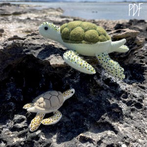 Può includere: Due tartarughe marine di feltro, una verde e una grigia, con dettagli perlati, sono sedute su una roccia grigia. La tartaruga più grande è in cima alla roccia, mentre la più piccola è sotto.