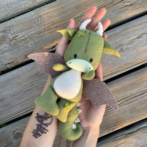 Puede incluir: Un peluche de dragón de fieltro verde hecho a mano con cuernos blancos, alas marrones y vientre blanco. El dragón tiene detalles cosidos y una expresión amigable. Un juguete suave y caprichoso para niños o coleccionistas.