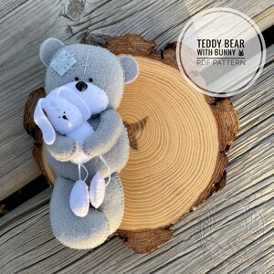 May include: A gray felt teddy bear hugging a white felt bunny. The teddy bear has a stitched patch on its head. The bunny has long ears and is holding two white felt dangling shapes. The text "TEDDY BEAR WITH BUNNY PDF PATTERN" is in a circle.