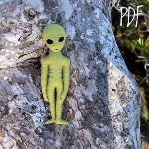 May include: A green felt alien doll with black eyes and a simple design. The doll is standing on a tree trunk.