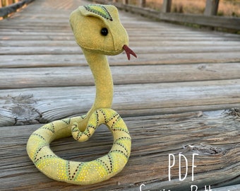 May include: A green and yellow felt snake sewing pattern. The pattern is for a plush snake with a patterned body and a small red tongue. The pattern is labeled "PDF Sewing Pattern".