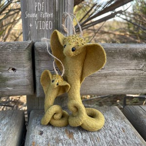 Puede incluir: Dos adornos de fieltro con forma de cobra en tonos verde oliva, con pequeños ojos negros y detalles de cuentas doradas. La cobra más grande tiene un cuerpo enrollado y un lazo para colgar. El texto "PDF Sewing Pattern + VIDEO" es visible.
