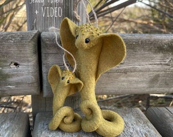 May include: Two felt cobra ornaments in shades of olive green, with small black eyes and gold bead accents. The larger cobra has a coiled body and a loop for hanging. The text "PDF Sewing Pattern + VIDEO" is visible.