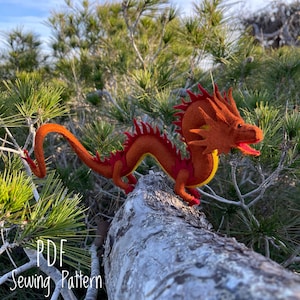 May include: A red felt dragon with yellow accents and a spiky mane, perched on a tree branch. The dragon is a sewing pattern, as indicated by the text "PDF Sewing Pattern" at the bottom of the image.