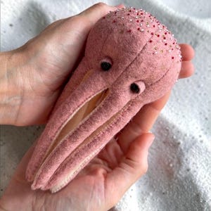 May include: A pink felt octopus toy with black button eyes and beaded detailing on its head. The octopus has three tentacles and is held in open hands. The background is a white, textured surface.