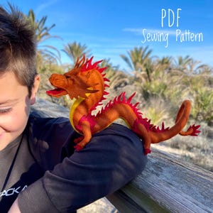 Peut inclure: Un jouet en forme de dragon en feutre rouge et jaune avec une longue queue et une crinière hérissée. Le dragon est assis sur un poteau de clôture en bois. Le texte "PDF Sewing Pattern" est visible dans le coin supérieur droit de l'image.