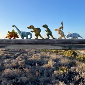 May include: Six colourful felt dinosaur toys, including a Stegosaurus, Brachiosaurus, and Triceratops, are arranged on a wooden fence. The dinosaurs are various colours, such as brown, blue, and green, against a clear blue sky.