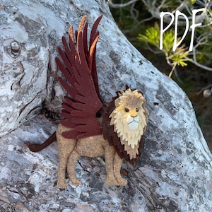 Puede incluir: Un león de fieltro hecho a mano con grandes alas de color rojo oscuro. El león tiene un cuerpo color canela, una melena marrón y una cara color crema. La palabra "PDF" está escrita en blanco en la esquina superior derecha. Artesanía.
